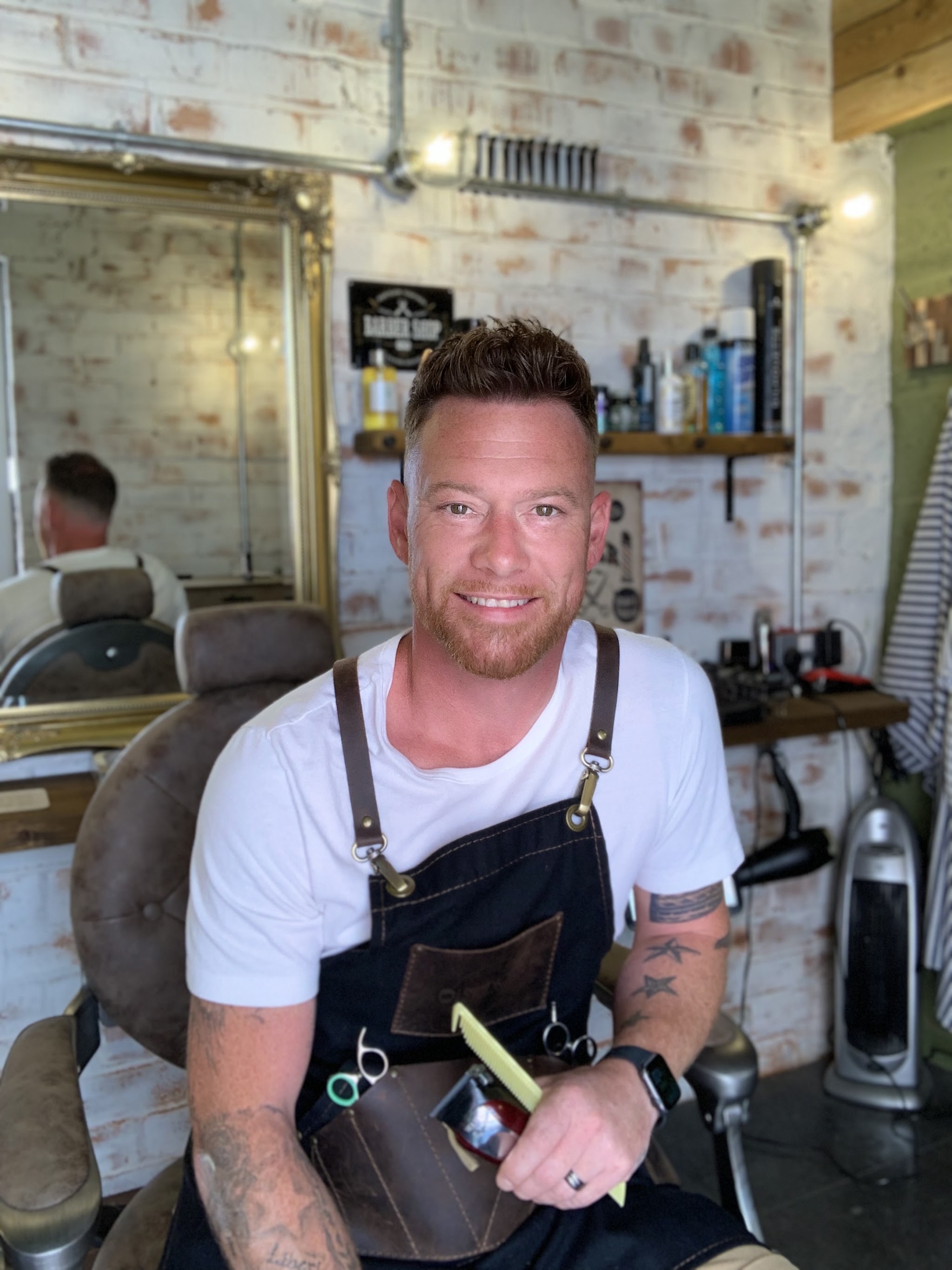 Dion White, the barber at Studio G, sitting in his barber's chair with scissors and a comb in his apron